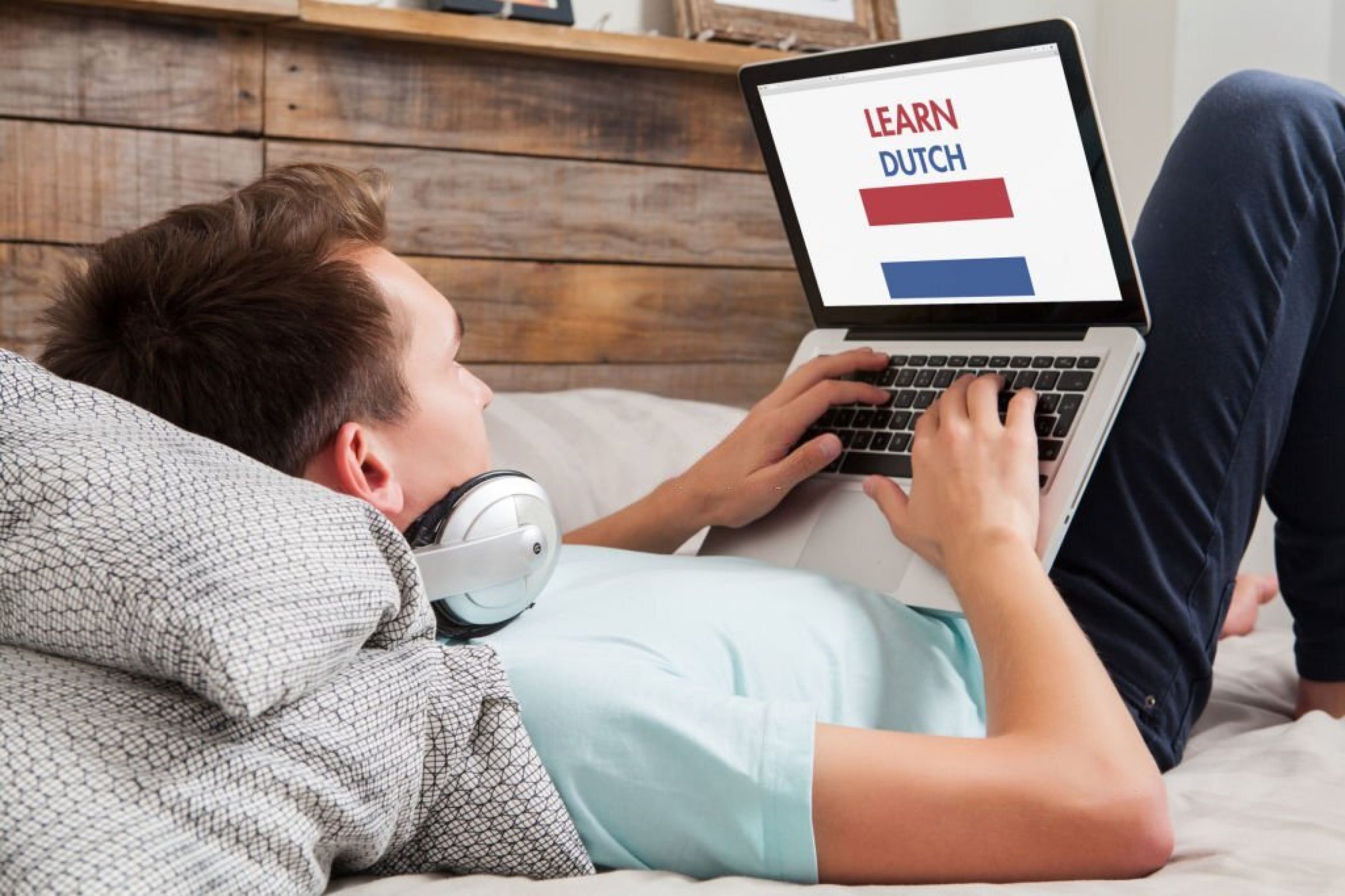 Man lying down on the bed at home while uses a laptop to learn Dutch language by internet.